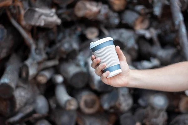 Testere kütüklerinin arka planında hazır kahve olan bir el. Çevredeki hasarın arka planında plastik bardak var. Gezegeni kurtar, plastik olmasın..
