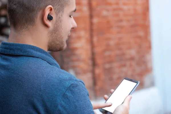 Young man listen music by wireless headphones and hold smartphone with white screen. Mock up for music album design cover or mobile application or web site. Back view. Keeping up with technology person.