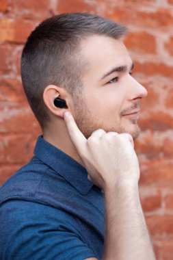 Genç yakışıklı adam müzik dinliyor ve kulağındaki kablosuz kulaklığı parmağıyla işaret ediyor. Bluetooth kulaklıklı modern bir adam. Yakın plan portre. Dikey