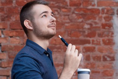 Bir elinde elektronik sigarayla genç yakışıklı bir adamın portresi ve diğer elinde kahve. Mavi tişörtlü çocuk öğle arasında sigara içiyor ve kahve içiyor. Kahve molası.. 