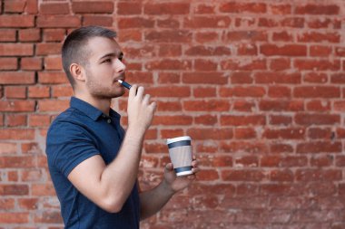 Bir elinde elektronik sigarayla genç yakışıklı bir adamın portresi ve diğer elinde kahve. Mavi tişörtlü çocuk öğle arasında sigara içiyor ve kahve içiyor. Kahve molası.. 
