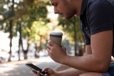 Genç adam akıllı telefonunun ekranına bakıyor ve parkta kahve içiyor. O, mavi bir tişört, mavi kot pantolon ve beyaz spor ayakkabı giyiyor. Tasarımınız için ekrandaki boşluk. Yatay