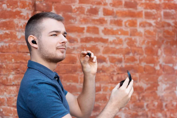 Young handsome man takes out a wireless earphone from a case and inserts it into his ear. Modern man listening music by bluetooth earphones. Keeping up with technology person.