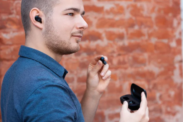 Young handsome man takes out a wireless earphone from a case and inserts it into his ear. Modern man listening music by bluetooth earphones. Keeping up with technology person.