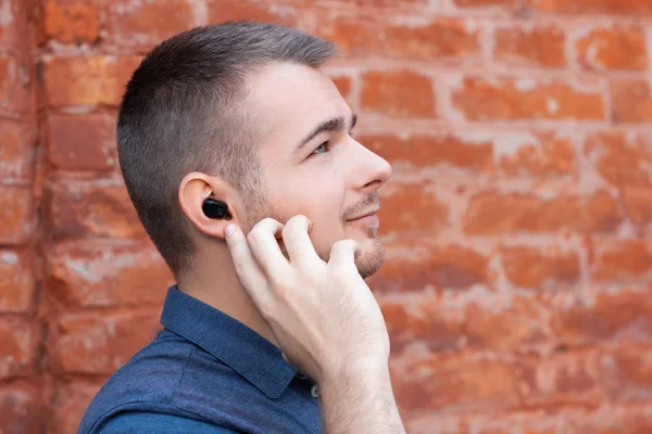 Genç yakışıklı adam müzik dinliyor ve kulağındaki kablosuz kulaklığı parmağıyla işaret ediyor. Bluetooth kulaklıklı modern bir adam. Yakın plan portre.