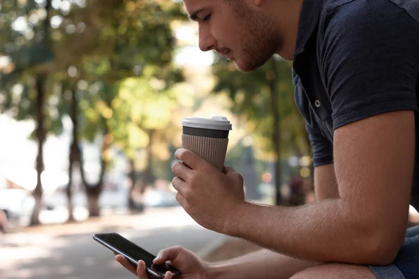 Young man is looking at the screen of his smartphone and drinking coffee in the park. He is wearing a blue t-shirt, blue jeans and white sneakers. Empty space on the screen for your design. Horizontal