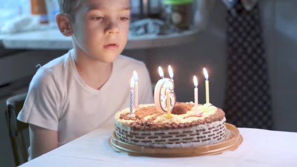 白いTシャツを着た6歳の白人の少年がキッチンのテーブルに座り 誕生日ケーキにろうそくを吹き 願い事をします パイの6番の形をしたキャンドル 家でお祝い — ストック動画