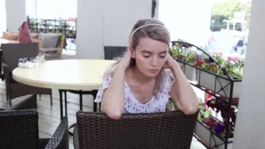 Kafe verandasında hasır sandalyede oturan ve poz veren beyaz elbiseli bir kız. Mavi gözlü güzel bir bayanın romantik videosu. Düşünceli bakışlar, yavaş şehvetli hareketler