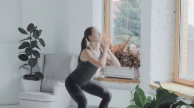 Spor giyimli esmer genç kız evde spor egzersizleri yapıyor. Çömeliyor.