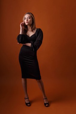 Siyah elbise ve turuncu arka planda poz veren yüksek topuklu ayakkabılar giyen zarif bir manken.