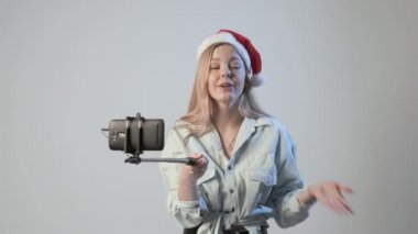 Noel Baba şapkalı genç kız selfie çubuğuna bağlı akıllı telefonuyla video kaydediyor.