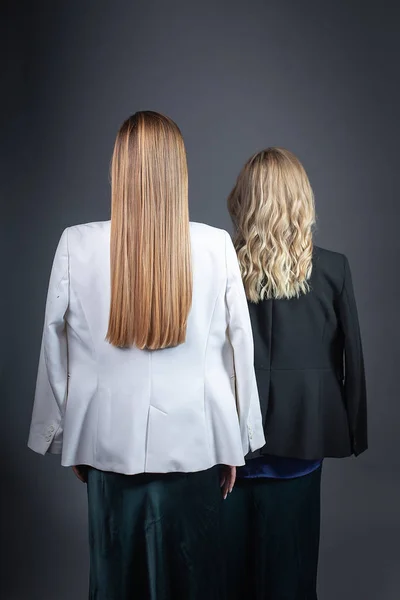 two girls with long blond hair stand against wall in strict suits, rear view