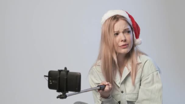 Молода дівчина в капелюсі Санта записує відео зі смартфоном, прикріпленим до палички селфі — стокове відео