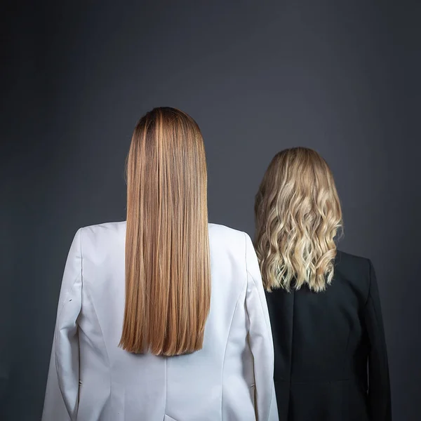 Two girls with long blond hair stand against wall in strict suits, rear view — Stock Photo, Image