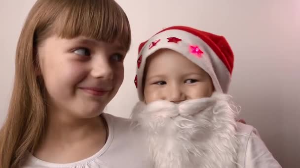 Zus en broer met kunstmatige baard in Santa Claus hoed glimlach, samen lachen — Stockvideo