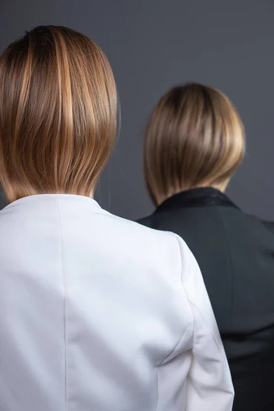 two girls with tucked blond hair stand against wall in strict suits, rear view