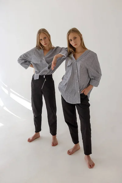 Dos hermanas gemelas muy jóvenes con el pelo largo y rubio posando sobre fondo blanco en ropa de gran tamaño. Sesión de fotos de moda —  Fotos de Stock