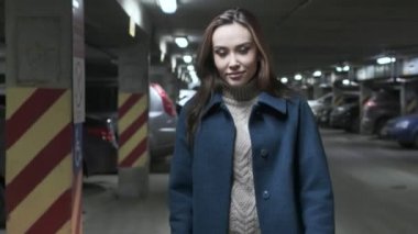 stylish smiling cute asian girl in coat walks at underground parking along cars