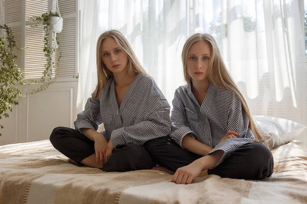 Симпатичные сестры-близнецы в джинсах и полосатых рубашках с длинными волосами, сидящие на диване — стоковое фото