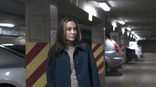 Elegant omtänksam asiatisk flicka i pälspromenader på underjordisk parkering längs bilar — Stockvideo