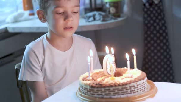 台所のテーブルに座って誕生日ケーキにろうそくを吹いて願い事をする男の子 — ストック動画