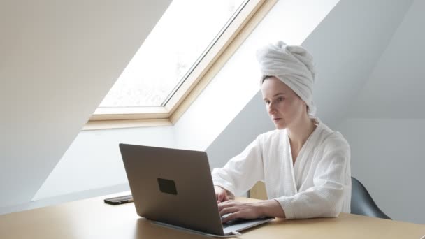 Jonge vrouw in witte badjas en handdoek op het hoofd werkt op afstand thuis op laptop — Stockvideo