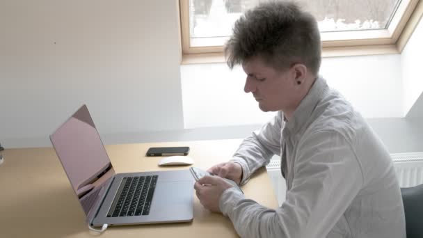 Hombre se sienta en la mesa con el ordenador, mira fijamente al teléfono inteligente. trabajar en casa — Vídeos de Stock