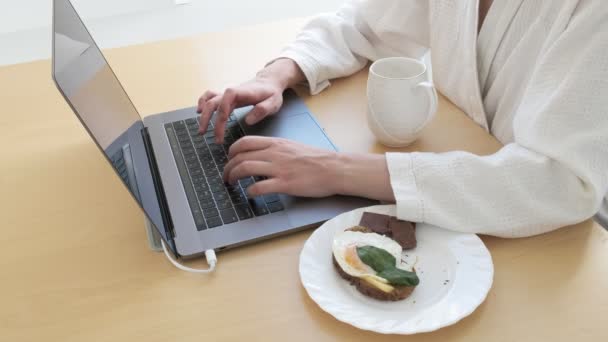 Dziewczyna pijąca kawę, jedząca śniadanie podczas pisania na klawiaturze laptopa, zbliżenie — Wideo stockowe