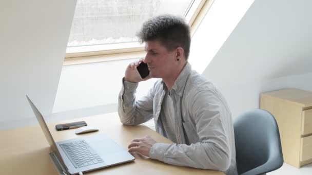 Hombre trabaja en la computadora, habla a través de teléfono inteligente. trabajar en casa en cuarentena — Vídeos de Stock