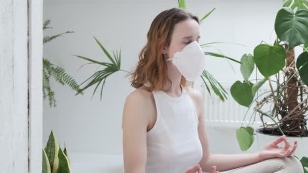 Een meisje met een beschermend masker zit in lotushouding mediterend. thuisblijven concept — Stockvideo