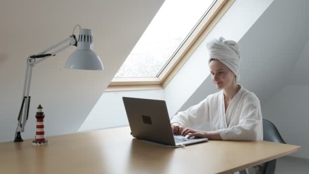 Junge Frau in weißem Bademantel und Handtuch auf dem Kopf arbeitet zu Hause am Laptop — Stockvideo