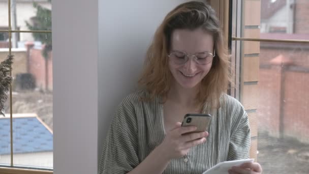 Meisje met bril zit op vensterbank, chats via smartphone. Online communicatie — Stockvideo