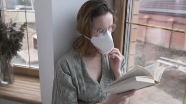 Koruyucu maskeli genç bir kız, gözlüklü, pencere kenarında oturuyor, roman okuyor.