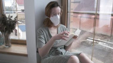 Koruyucu maskeli genç bir kız, gözlüklü, pencere kenarında oturuyor, roman okuyor.