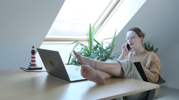 快乐的高加索女人下班后一边说着带腿的智能手机，一边放松地坐在桌子上 — 图库视频影像