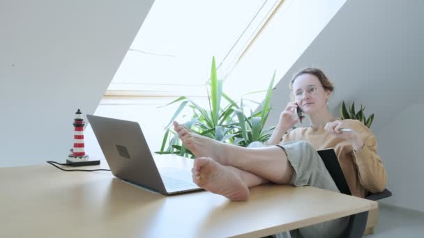 Щаслива біла жінка розмовляє смартфоном з ногами на столі, розслабляючись після роботи — стокове відео