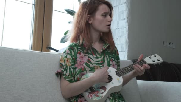 Menina bonita sentada no sofá toca guitarra de brinquedo, tolos ao redor, canta música, sorrisos — Vídeo de Stock