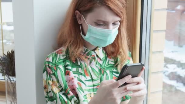 Ragazza in maschera protettiva sms in smartphone sul davanzale della finestra. auto isolamento — Video Stock