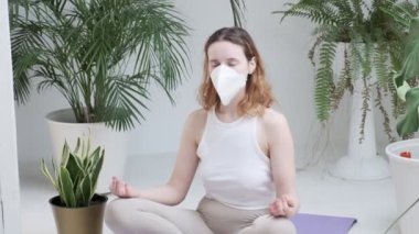 Spor giyimli bir kız, koruyucu maskeli parlak odada meditasyon yapıyor.