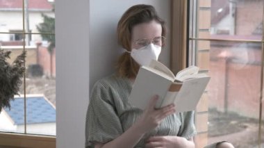 Koruyucu maskeli, gözlüklü, pencere kenarında oturan, şiir kitabı okuyan genç bir kız.