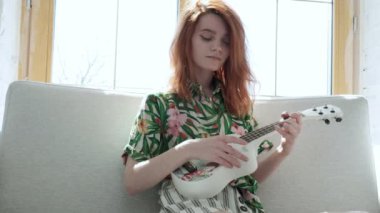 Çiçekli tişörtlü kız güneşli bir günde parlak bir odada, koltukta gitar çalıyor.