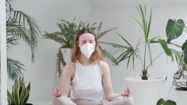 Spor giyimli bir kız, koruyucu maskeli parlak odada meditasyon yapıyor.