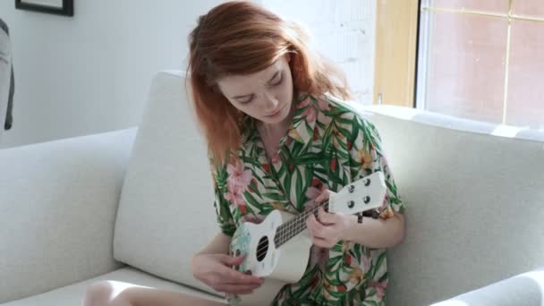 Девушка в цветущей рубашке, играющая на укулеле на диване в светлой комнате в солнечный день — стоковое видео