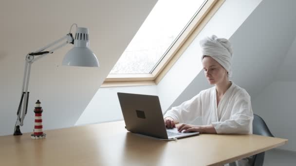 Молода жінка в білому халаті і рушник на голові дистанційно працює вдома на ноутбуці — стокове відео