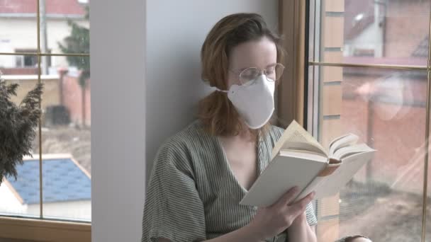 Νεαρή κοπέλα με προστατευτική μάσκα, γυαλιά κάθεται στο περβάζι του παραθύρου, διαβάζει ποιήματα βιβλίο — Αρχείο Βίντεο