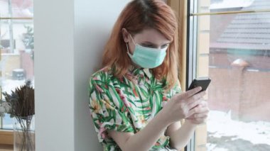 Koruyucu maskeli kız pencere eşiğinde akıllı telefondan sohbet ediyor. kişisel izolasyon