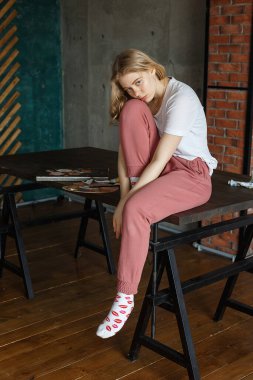 Evde kalmak, yaratıcı kriz kavramı: üzgün, genç, sarışın, güzel kadın sanatçı, resim, fırça ve paletle birlikte sehpanın yanında oturuyor. COVID-19 salgını sırasında boyama işlemi kendini izole etme karantinası