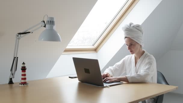 Junge Frau in weißem Bademantel und Handtuch auf dem Kopf arbeitet zu Hause am Laptop — Stockvideo