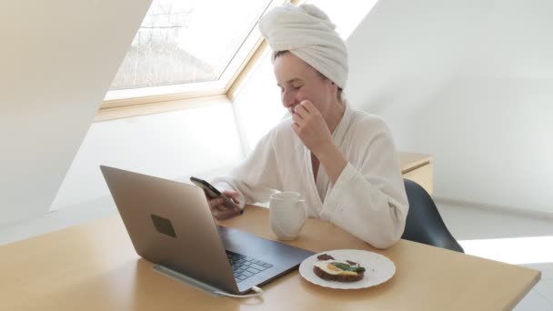 Mulher freelance em roupão de banho na mesa, fala smartphone, tipos no teclado do laptop — Vídeo de Stock