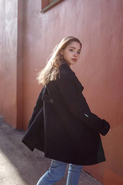 화창 한 날검은 코트를 입고 검은 코트를 입은 아름다운 소녀가 빨간 벽에 몸을 돌립니다. 거리 양식 초상화 — 스톡 사진